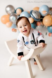 space cake smash. a one year old boy in a space onsie is in milwaukee photography studio for birthday pictures. he's in front of a white backdrop with balloons in shades of blue, silver, and beige with string lights and planets on them.