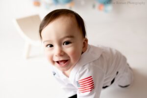 space cake smash. one year old boy in milwaukee photography studio. close up image of his face smiling, with cute little teeth. boy has a space onsie on, and the backdrop is white with a balloon garland.