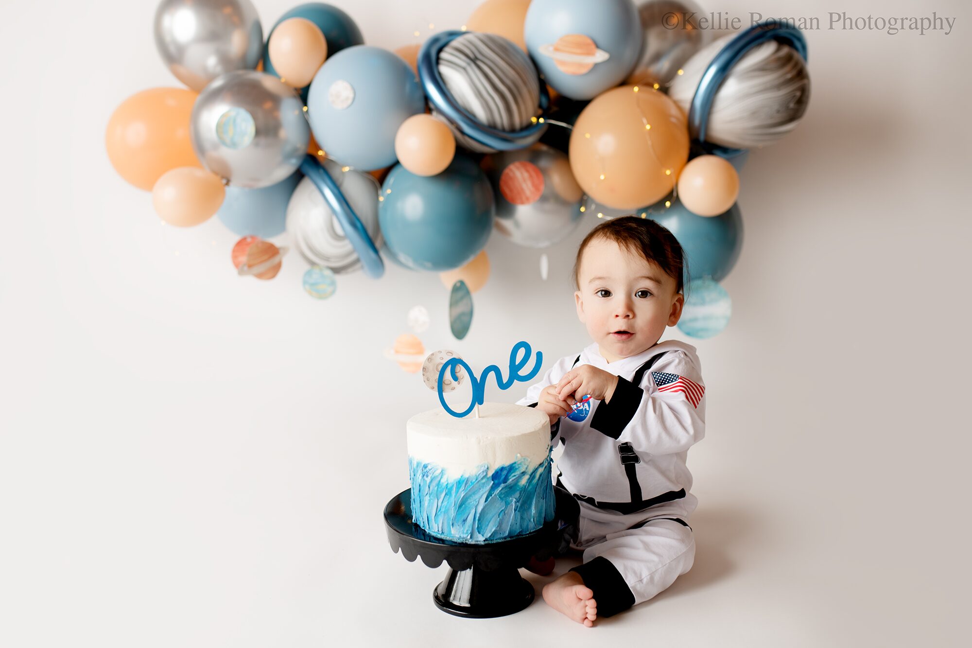space cake smash. one year old boy in greendale photography studio. boy is wearing an astronaut outfit sitting behind a blue and white cake with a blue one on the top. the floor and backdrop is white with a blue, silver, and beige balloon garland. there are small lights in the balloons and space cut outs.
