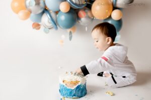 space cake smash. one year old boy wearing an astronaut outfit is sitting on white backdrop and floor with a silver blue and beige balloon garland behind him.