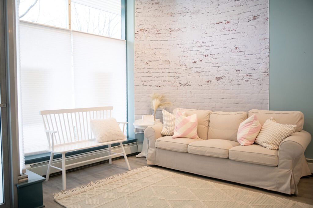 new milwaukee photography studio. the image is the client seating in bayview photo studio. there are two big windows with shades, a white bench and cream couch. there are pink and cream pillows on the couch with a cream and teal rug. the wall is blue and has a cream brick backdrop hanging. 