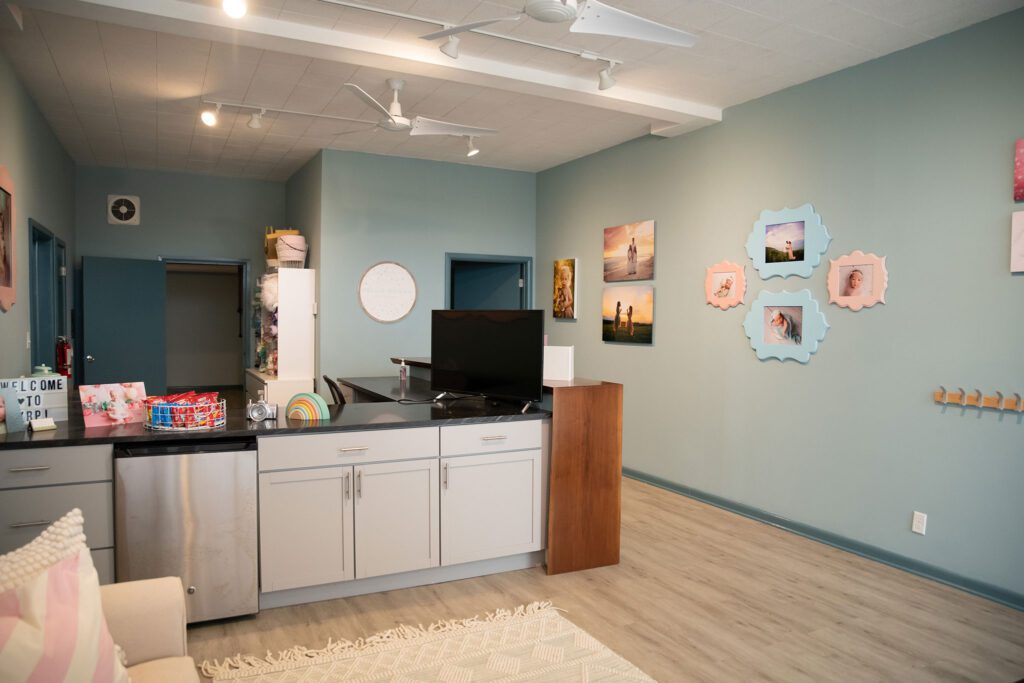 new milwaukee photography studio. the image is inside a bayview photo studio the walls are light blue with lots of client pictures hanging in pastel frames. the floors are light beige, and there is a large built in desk with light grey cabinets and a black countertop. 
