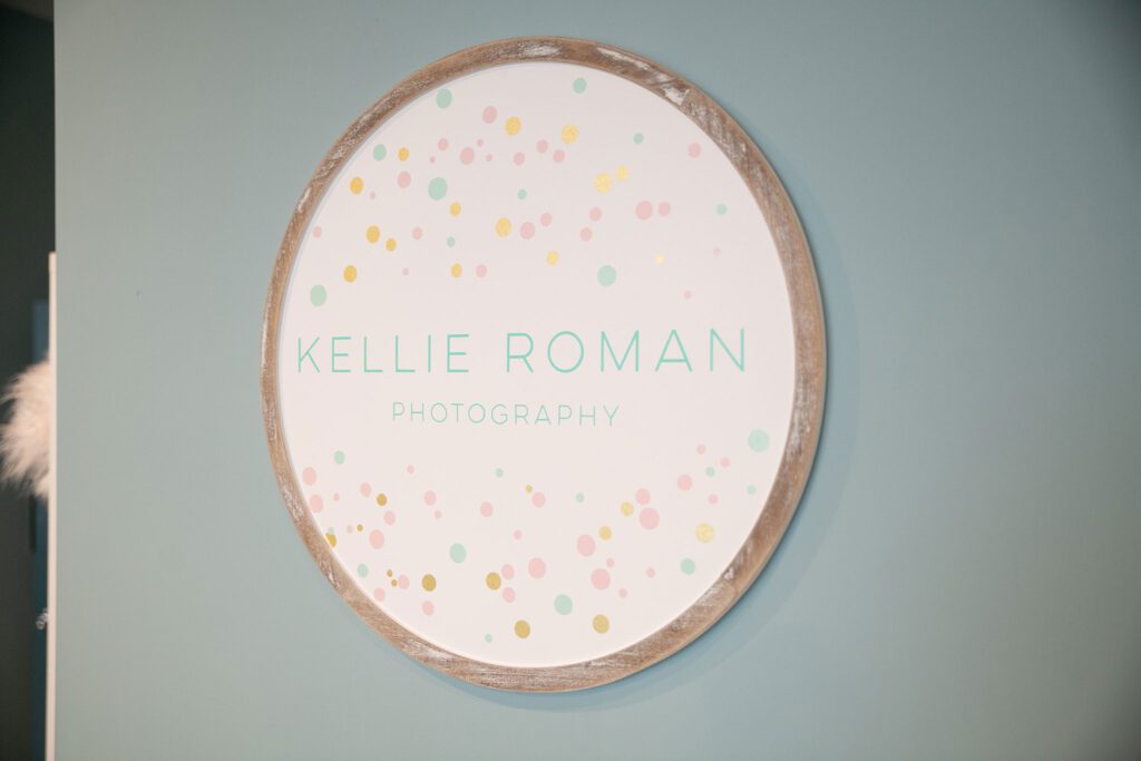  a close up of a wood round sign with kellie roman photography logo. the logo is light pink pastel, teal, and gold. the sign is hanging on a blue wall. 