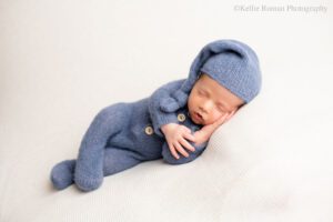 milwaukee newborn pictures near me. a newborn boy is onto of cream fabric sleeping on his side. he has his hand under his head. he's wearing a blue footed romper with a matching blue hat.