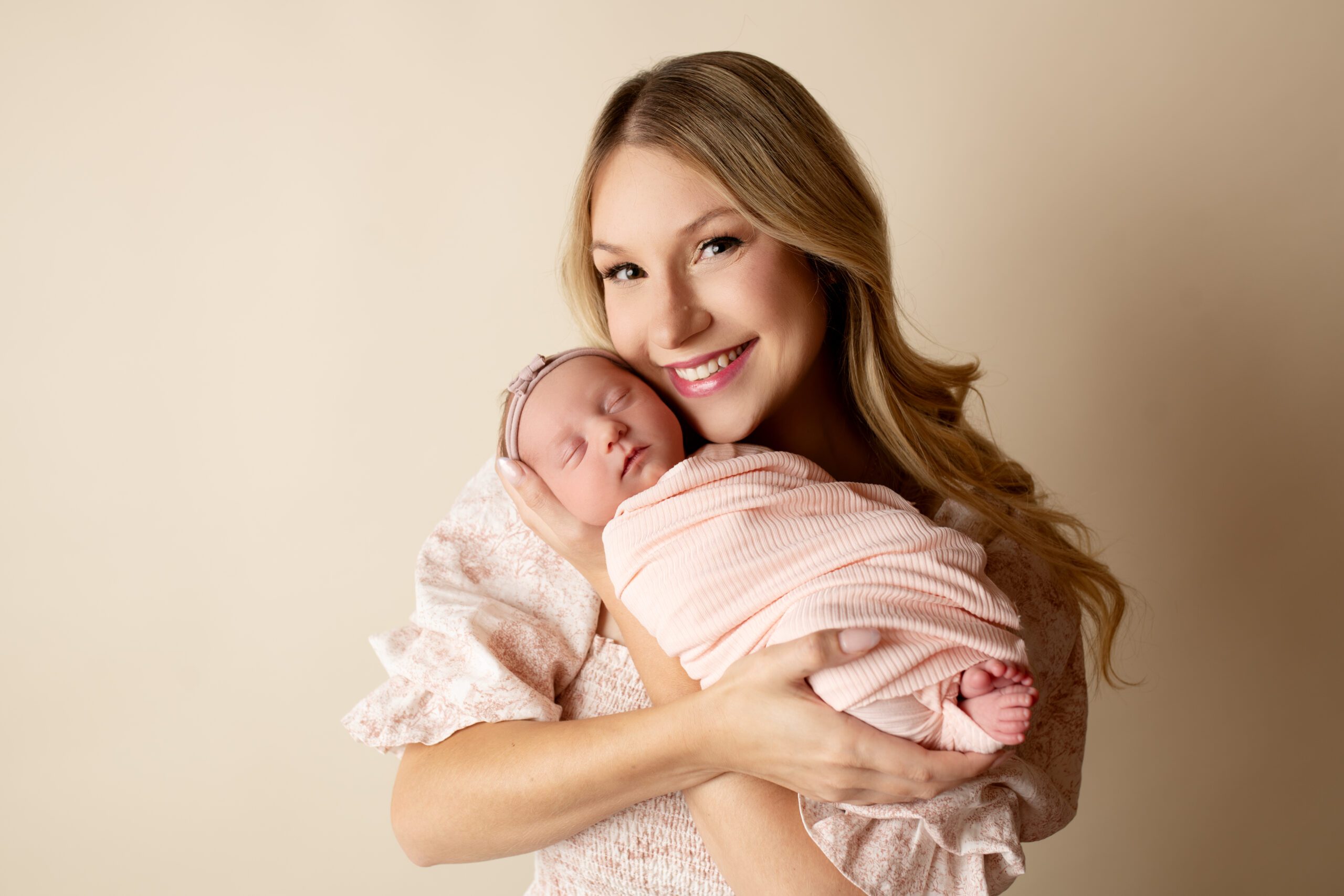 a new mother is holding her newborn daughter up her her face and is looking and smiling at the camera. baby is sleeping and swaddled in a light pink swaddle.
