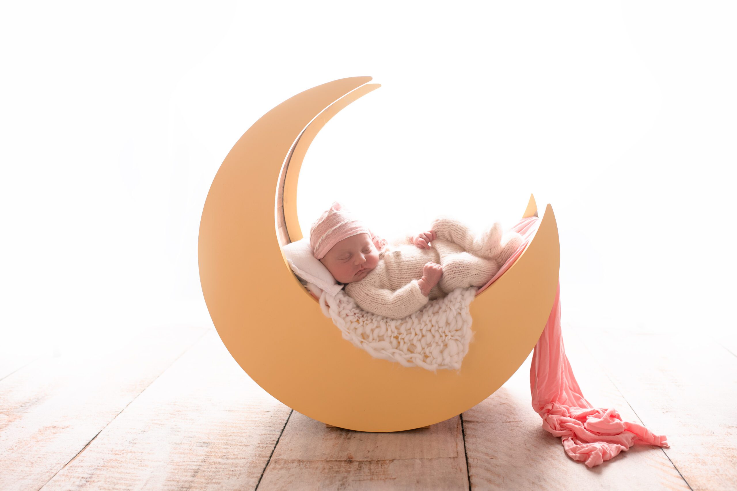 newborn girl is laying on her back onto of a wood yellow moon newborn prop. the moon has a white pillow for the newborns head and a white knit fabric as well as a long pink fabric hanging off the side. newborn is in white knit footed romper with a pink and white striped hat. lots of bright light behind the baby
