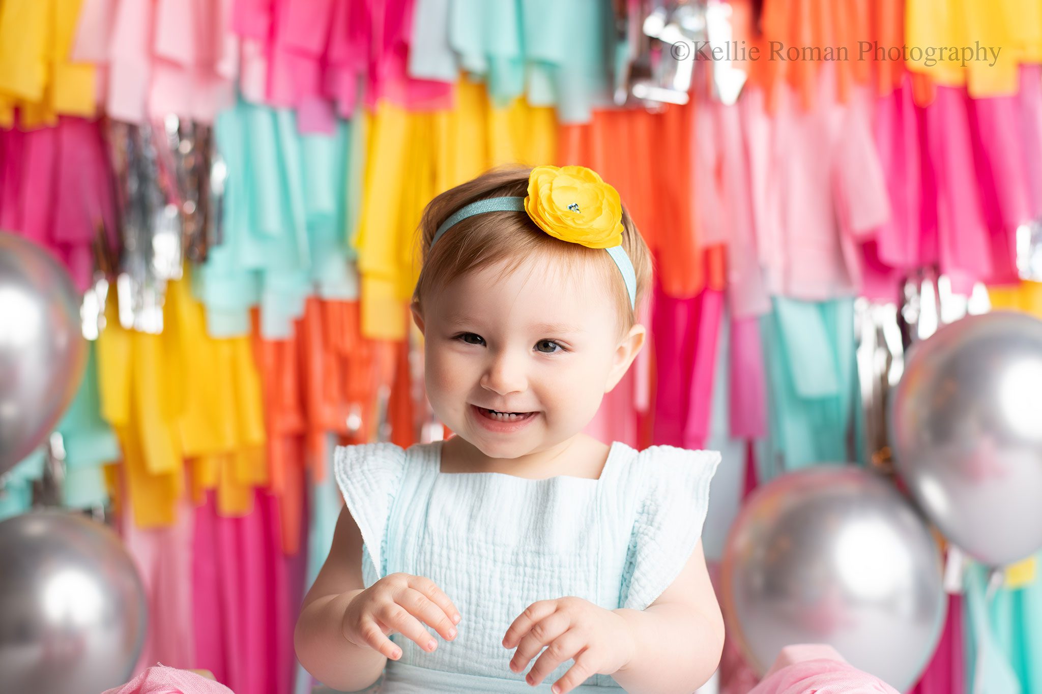 one year old is smiling showing her teeth. she has light blue romper and matching headband on. the backdrop is very bright with lots of colors. blue pinks, oranges, yellows, and silvers.
