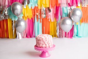 festive cake smash photo session in milwaukee. pink rosette cake is onto of a dark pink cake stand. the backdrop is hand made with lots of fringe colors of yellow, orange, teal, pink and silver. there are silver balloons.