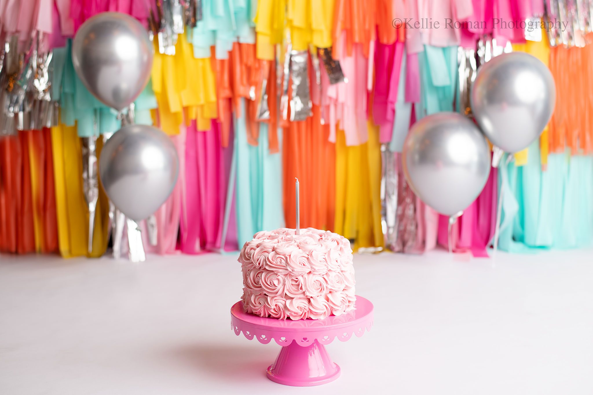 cake smash in milwaukee photo studio. light pink cake full of rosettes is resting on dark pink cake stand. the floor is white paper, and the backdrop has tons of bright colors. light pink, teal, yellow, orange and silver.
