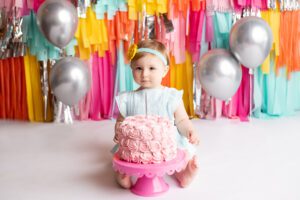 fringe cake smash milwaukee. a one year old girl sitting behind pink frosted cake. she has a blue romper on with a matching headband. she's looking at the camera and not smiling. the backdrop is fun bright colors of pink, yellow, orange, teal, and silver.