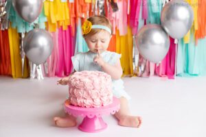 fringe cake smash milwaukee. a one year old girl sitting behind pink frosted cake. she has a blue romper on with a matching headband. she's has her finger in her mouth and is looking down. the backdrop is fun bright colors of pink, yellow, orange, teal, and silver.