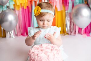fringe cake smash milwaukee. a one year old girl sitting behind pink frosted cake. she has a blue romper on with a matching headband. she's looking down at the cake. the backdrop is fun bright colors of pink, yellow, orange, teal, and silver.
