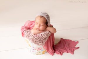 stunning newborn session. newborn baby girl is sleeping and posed in a flower bucket. she has her chin resting on her arms with a pink mohair bonnet on showing off her dark brown hair. the bucket has pink and mauve knit and fabric layers in it.