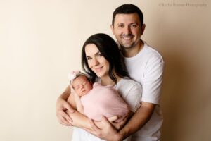 stunning newborn session. a mother and father with their newborn daughter in milwaukee photo studio. mother is holding newborn girl who is in a pink swaddle wrap with flower headband. dad is standing behind and holding onto both the mother and baby girl. parents have white shirts on.