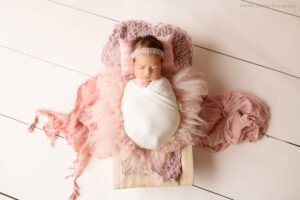 stunning newborn session. newborn girl is swaddled in cream wrap and is sleeping. she's onto of a wood crate filled with different pink furs and fabrics. she has one hand sticking out of the wrap and a bead headband on,.