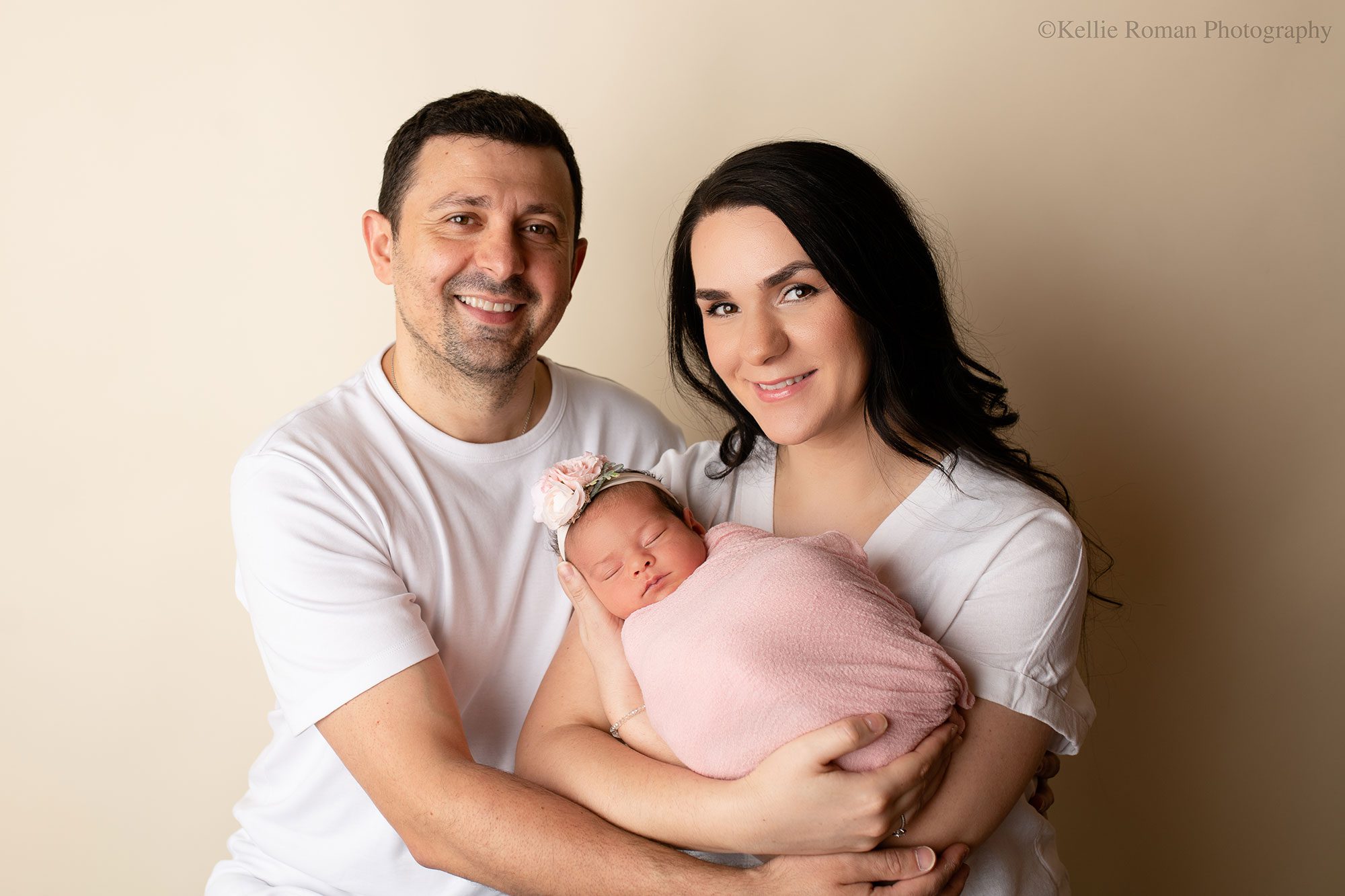 stunning newborn session. 
family of three in milwaukee photo studio. new parents are smiling at the camera. both parents are wearing white shirts. mother is holding newborn girl who has on a light pink swaddle around her and a light pink flower headband.

