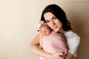 stunning newborn session. a beautiful new mother is holding her newborn daughter while smiling and looking at the camera. the mom has a white shirt on. baby girl is swaddled in a light pink wrap with a pink floral headband on.