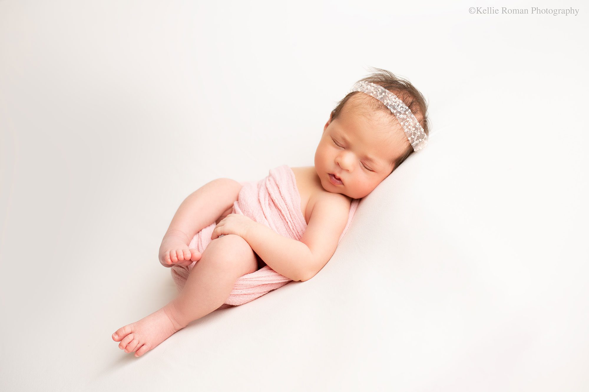 stunning newborn session. newborn girl in milwaukee photography studio is laying on her back asleep on cream fabric. she has a loose light pink swaddle around her, but her arms and legs are hanging out. she has a sequin light pink headband on. 

