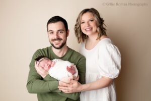 little newborn changes. family of three in milwaukee photo studio infront of cream backdrop. mom and dad have newborn baby, and dad is holding infant. dad has a green shirt on, mom has a cream dress on. newborn has cream wrap with her toes sticking out, and cream floral headband.