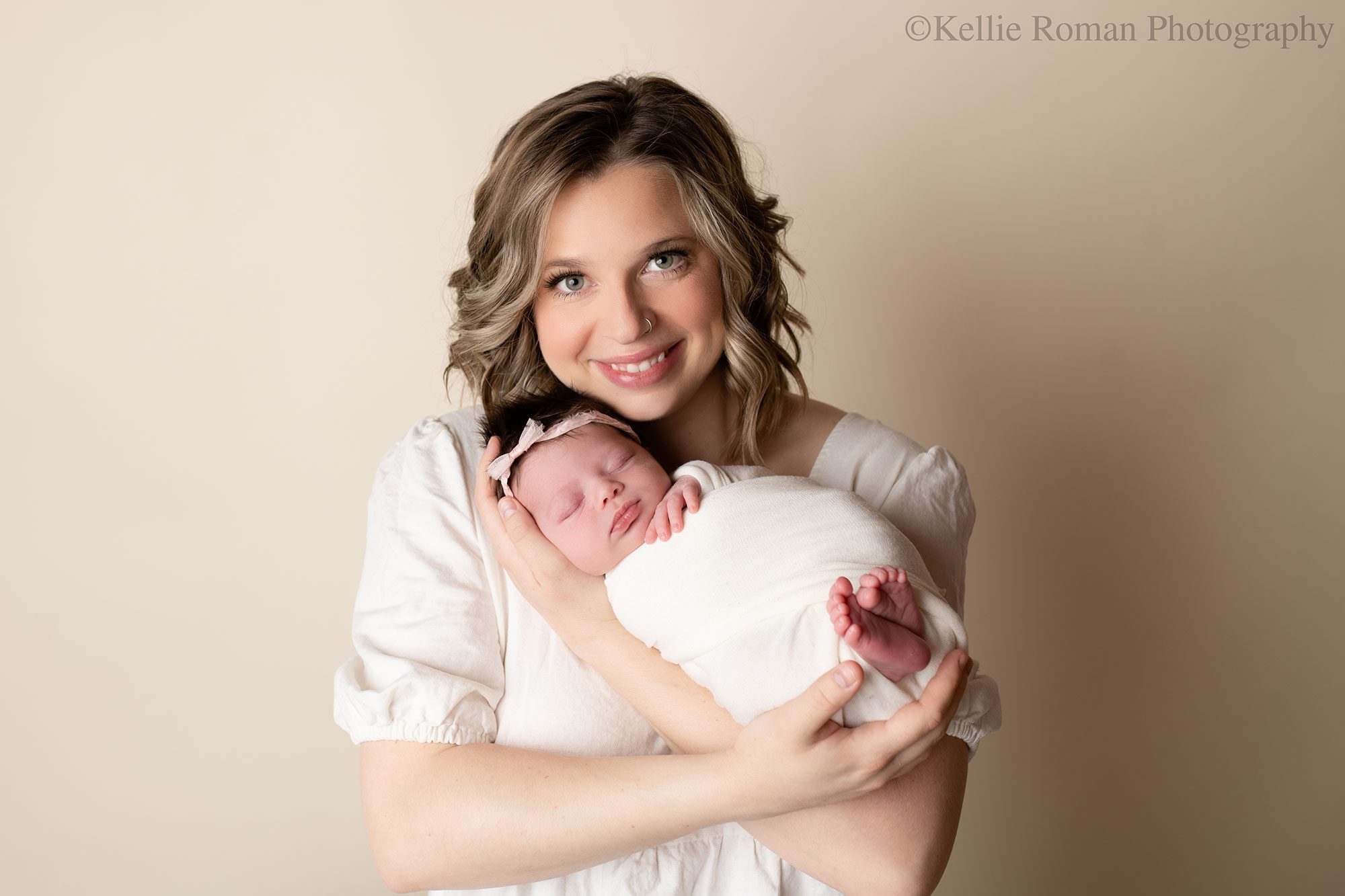 little newborn changes. a new mother is holding her daughter in milwaukee photographers studio. mother has a cream dress on and newborn is wrapped in cream swaddle with her toes and hand sticking out. she has a floral cream headband on. 

