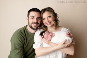little newborn changes. a husband and wife are in milwaukee photography studio is smiling at the camera. mom is holding her newborn daughter who's in a cream, swaddle with floral headband. mom has a cream dress on and dad has a green shirt.