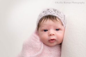 little newborn changes. newborn girl in milwaukee photo studio. she's swaddled in a light pink mohair fabric with matching bonnet. she has dark hair sticking out of the bonnet. she's wide eyed.