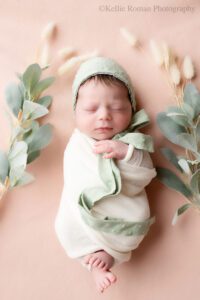 little newborn changes. newborn girl in mke photography studio. she's swaddled in a cream wrap with her toes and one hand sticking out. she has a green bonnet on, and is surrounded by flowers. she's laying onto of a light pink fabric.