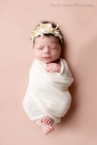 little newborn changes. milwaukee newborn photo studio with newborn baby swaddle in cream wrap with her toes sticking out. she has a floral cream headband on. her one hand Is sticking out of the top of the wrap. she's sleeping on her back onto of light pink fabric.