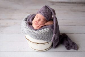newborn photography milwaukee. newborn girl sleeping in a white wood bucket that is filled with grey and purple layers. the baby has her chin resting on her arms with a dark purple sleepy cap on.