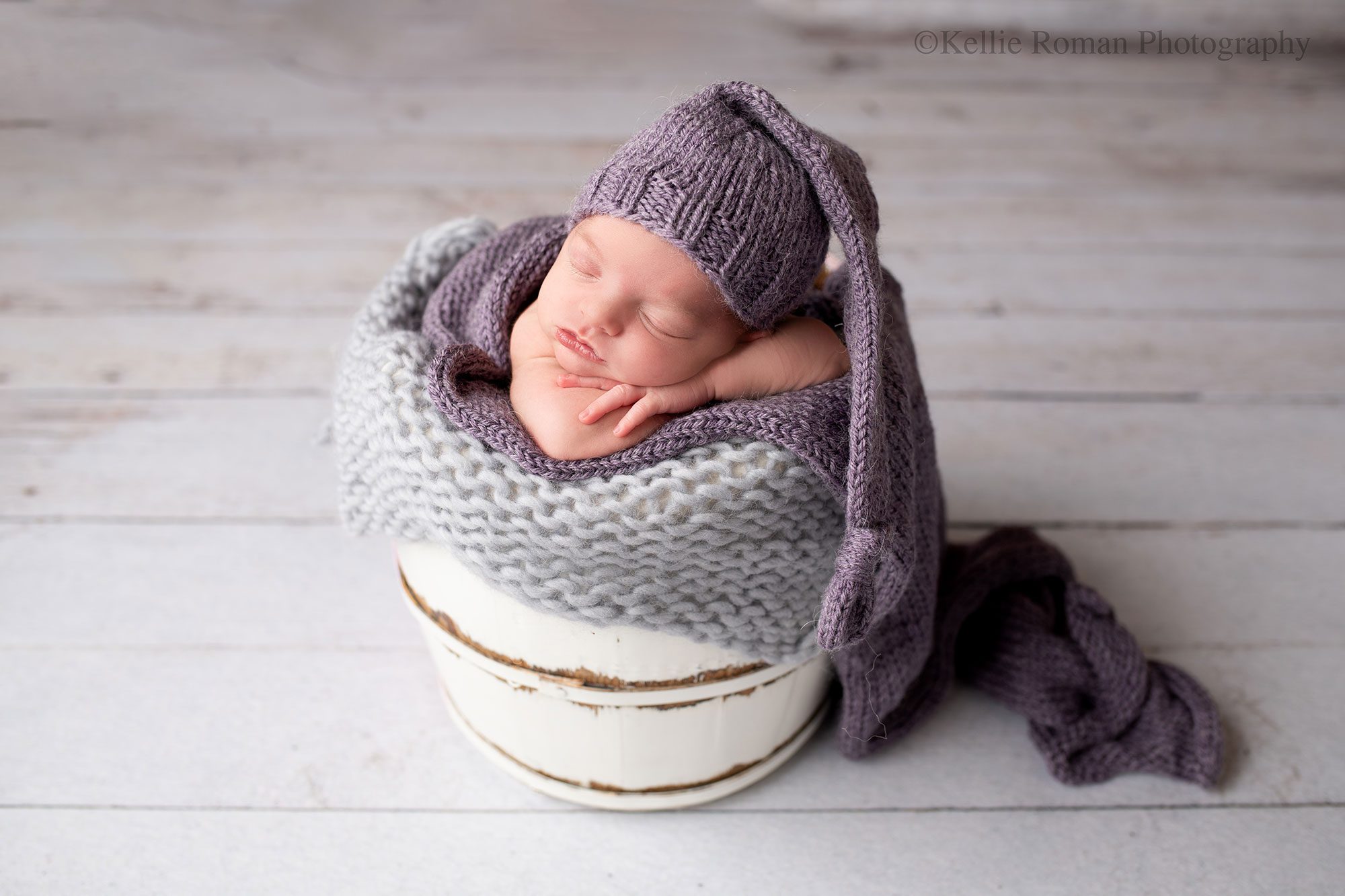 newborn photography milwaukee. a newborn baby girl is sleeping in a white wood bucket. baby has her chin resting on her arms and hands and is wearing a purple sleepy hat. the wood bucket has layers of grey and purple fabrics in it. 

