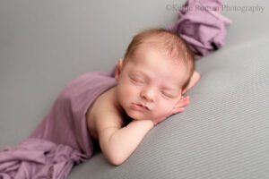 newborn photography milwaukee. newborn girl sleeping with her chin resting on her hands. she's laying onto of grey fabric with a deep purple fabric draped over her diaper.