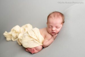 newborn photography milwaukee. newborn girl is laying on her belly on a light grey fabric. she has a light yellow swaddle wrap draped over her back and diaper. she has her hand resting under her cheek.