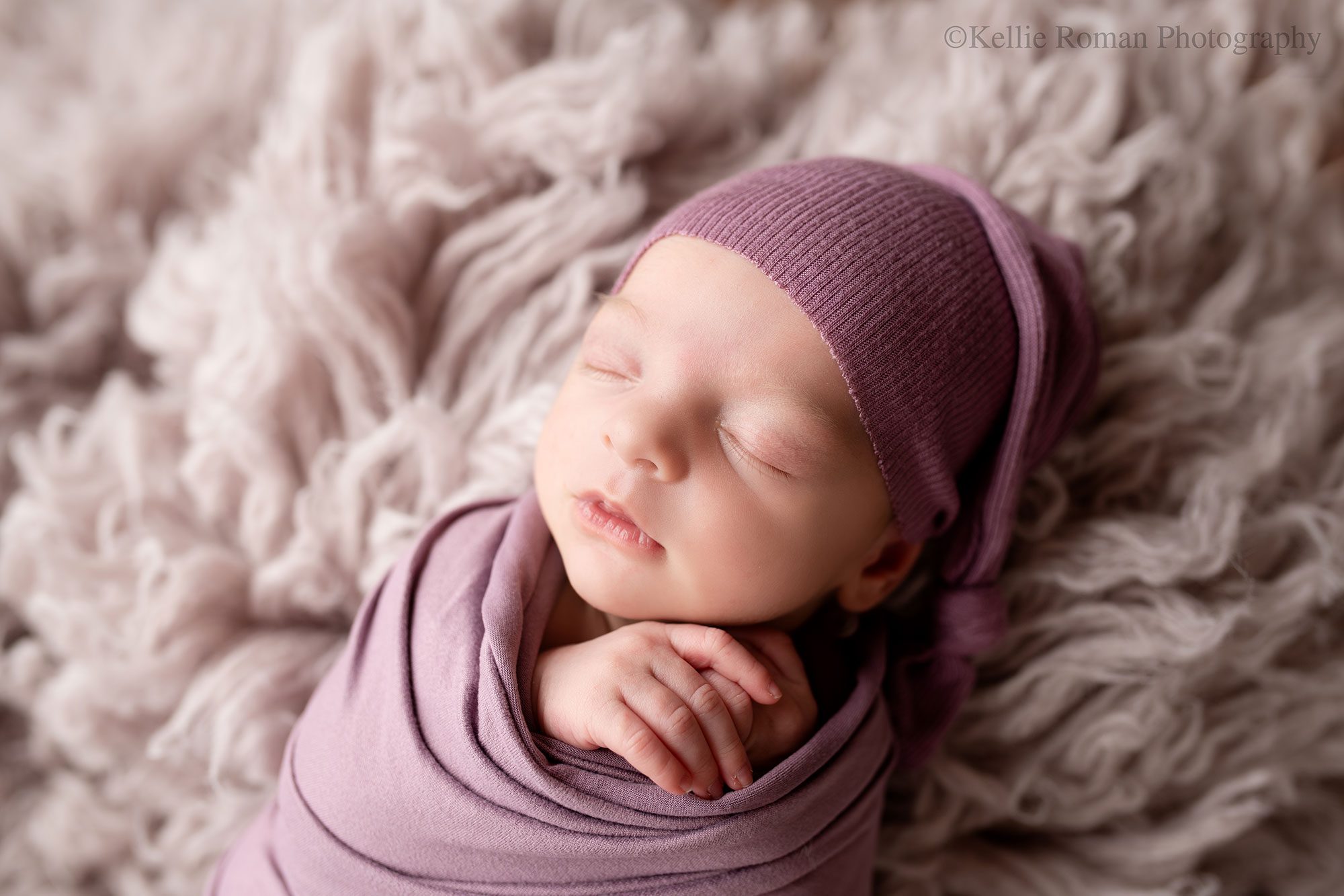 newborn photography milwaukee. newborn girl is in a dark purple swaddle with matching purple sleepy had. she's sleeping with her hands sticking out of the wrap onto of a purple fuzzy rug. 
