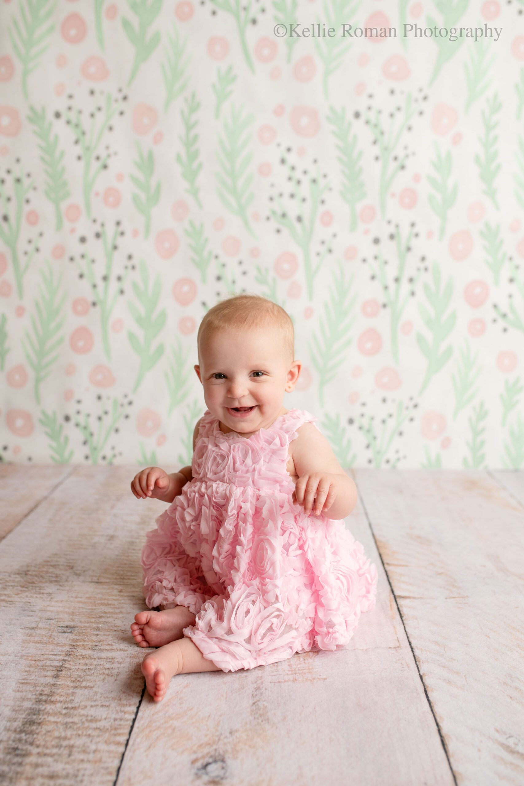 milwaukee baby photography. 6 month old baby sitting on her own wearing a bubble gum pink dress and smiling. the backdrop has green pink and grey floral pattern. baby is barefoot
