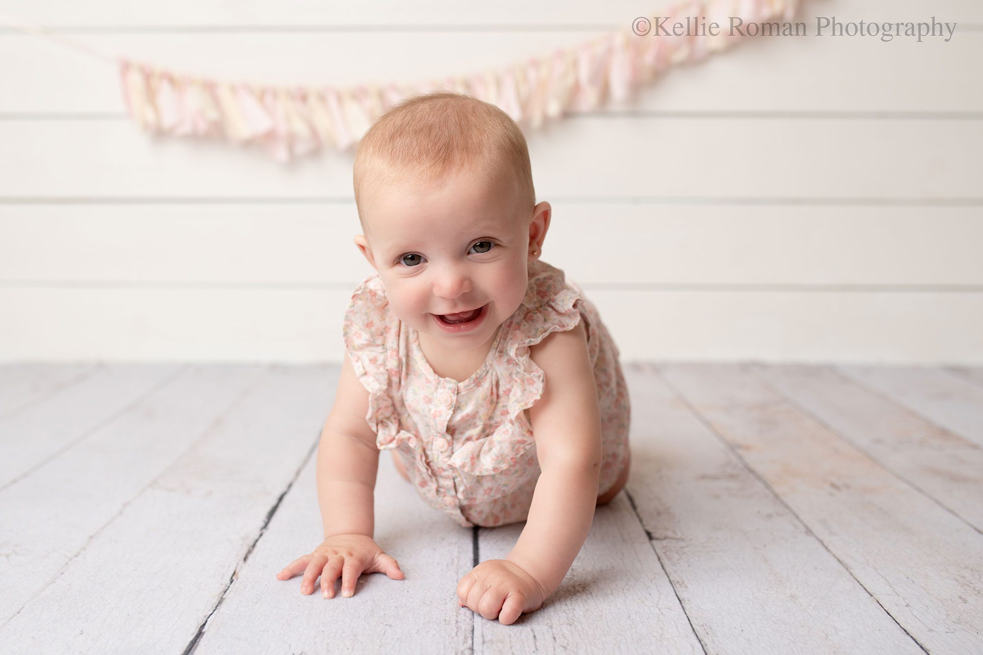 milwaukee baby photography. six month old baby girl is on her hands and knees crawling forward and smiling big. she's looking right at the camera and has a light pink and cream flower romper on. the backdrop is white wood with a pink and cream fabric banner.
