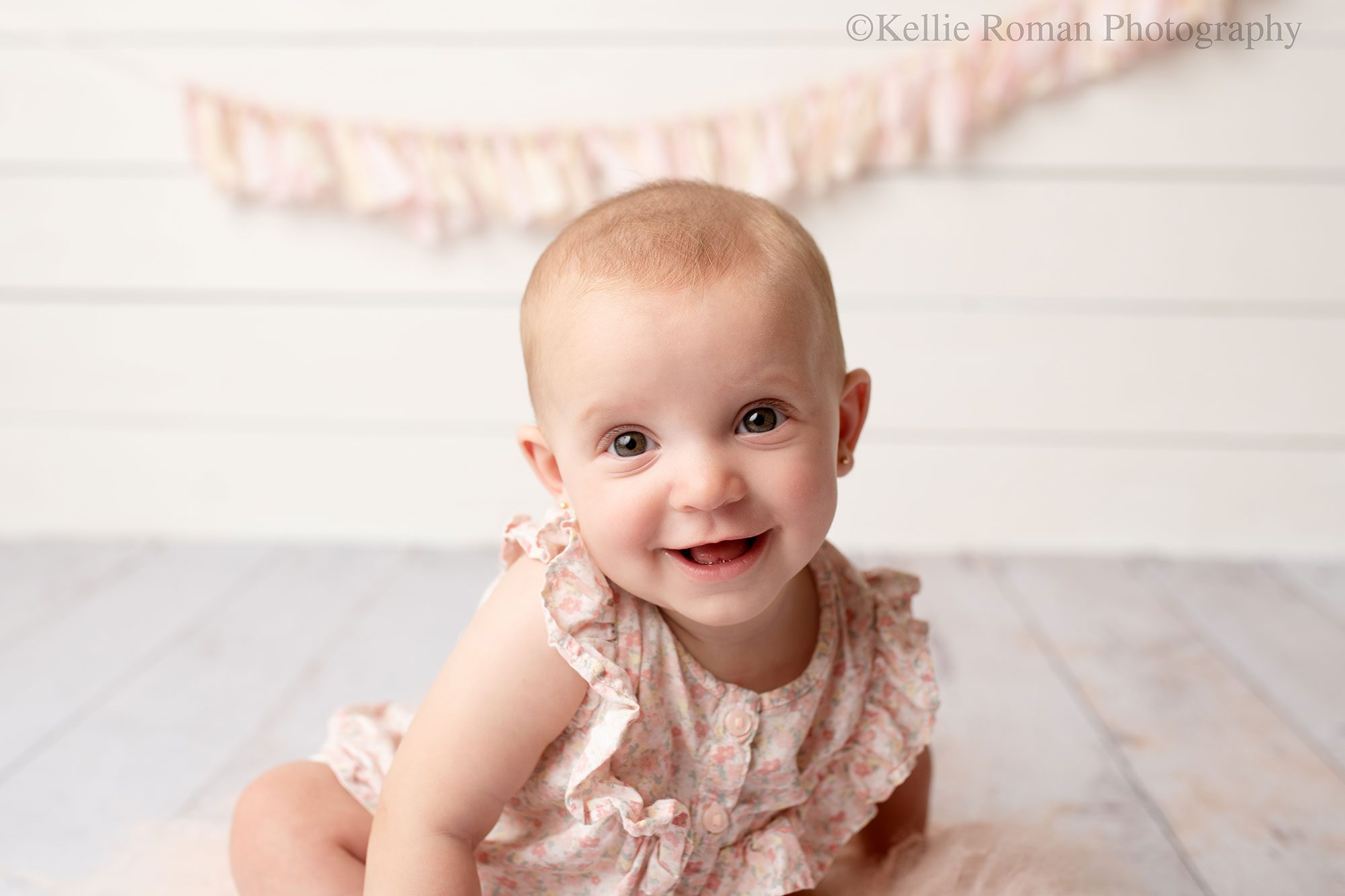 milwaukee baby photography. six month old baby girl is smiling at camera she has big brown eyes with a flower romper on. backdrop is white wood with a pink and cream fabric banner

