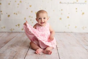 milwaukee baby photographer. six month old baby girl sitting up holding onto the edge of her dress. she's smiling big, while her toes are sticking out. the backdrop is cream with gold stars. her dress is bubble gum pink.