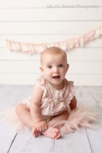 milwaukee baby photography. six month old baby girl sitting on white wood floor in front of a white wood backdrop with pink and cream banner. baby has a cream and light pink flower romper on. she has big brown eyes and is smiling.