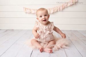 milwaukee baby photography. six month old girl is sitting on a wood floor onto of light pink fur. she has a light pink and cream floral romper on, and is smiling big at the camera. the backdrop is white wood with cream and light pink fabric banner.