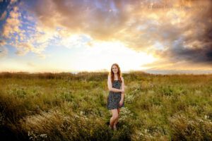 Senior photography milwaukee. high school senior girl standing in field of tall grass with stunning set set behind her. girl has curly red hair and is wearing a black dress with white polka dots.
