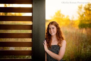 senior photography milwaukee. senior girl standing in park with tall grass and setting sun behind her. she's leaning against wood and metal wall, with her hand up on the wall. she has a black dress with white polka dots on, and long red hair.
