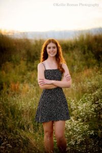 senior photography milwaukee. high school senior girl standing in tall green grass with black dress with polka dots. sun is setting behind her. the girl has red curly long hair.