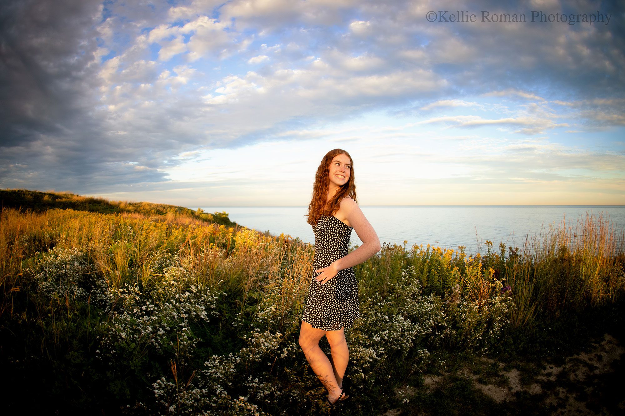 senior photography milwaukee. high school senior girl stand with her hand on her hip and is looking behind her. Lake Michigan and lots of tall grass is behind her. girl has red curly hair and a dress with white polka dots.