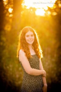 senior photography milwaukee. high school senior girl in milwaukee park with golden sun setting behind her through the leaves. she's has a black dress with white polka dots on it. she's holding her arm with her hand and smiling at camera.
