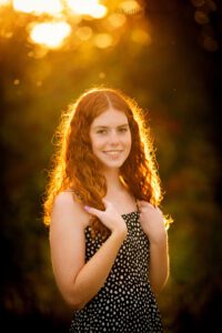 senior photography milwaukee. senior girl wearing black dress with white polka dots is standing with sun setting behind her. her red hair is light up with golden tones from sun, she's holding onto her hair.