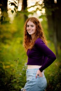 senior photography milwaukee. senior girl with long red curly hair in Milwaukee County park. she's standing turning behind her with hand in back jeans pocket. she has a purple shirt on.