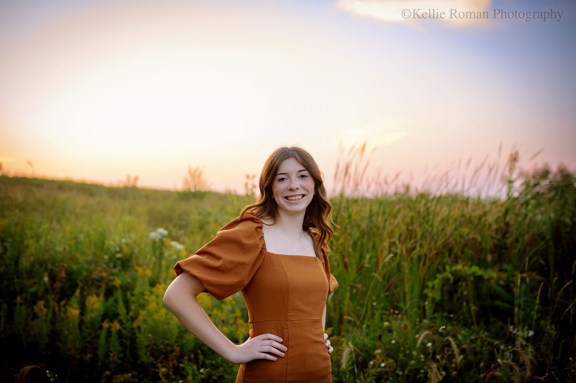 senior pictures in milwaukee. high school senior Muskego girl wearing a light brown dress standing in field of tall grass with sun setting behind her. girl has her hands on her hips.