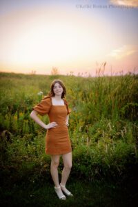 senior pictures in milwaukee. high school senior girl wearing a brown dress standing in tall grass in milwaukee park with sun setting behind her. she has her hands on her hips and wind is blowing through her hair.