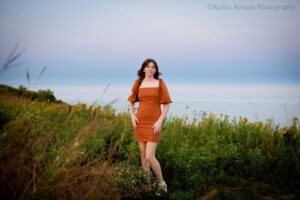 senior pictures in milwaukee. milwaukee high school senior girl wearing a brown short dress standing in grassy field in front of Lake Michigan. she has her hands on her hips and the wind is blowing through her hair.