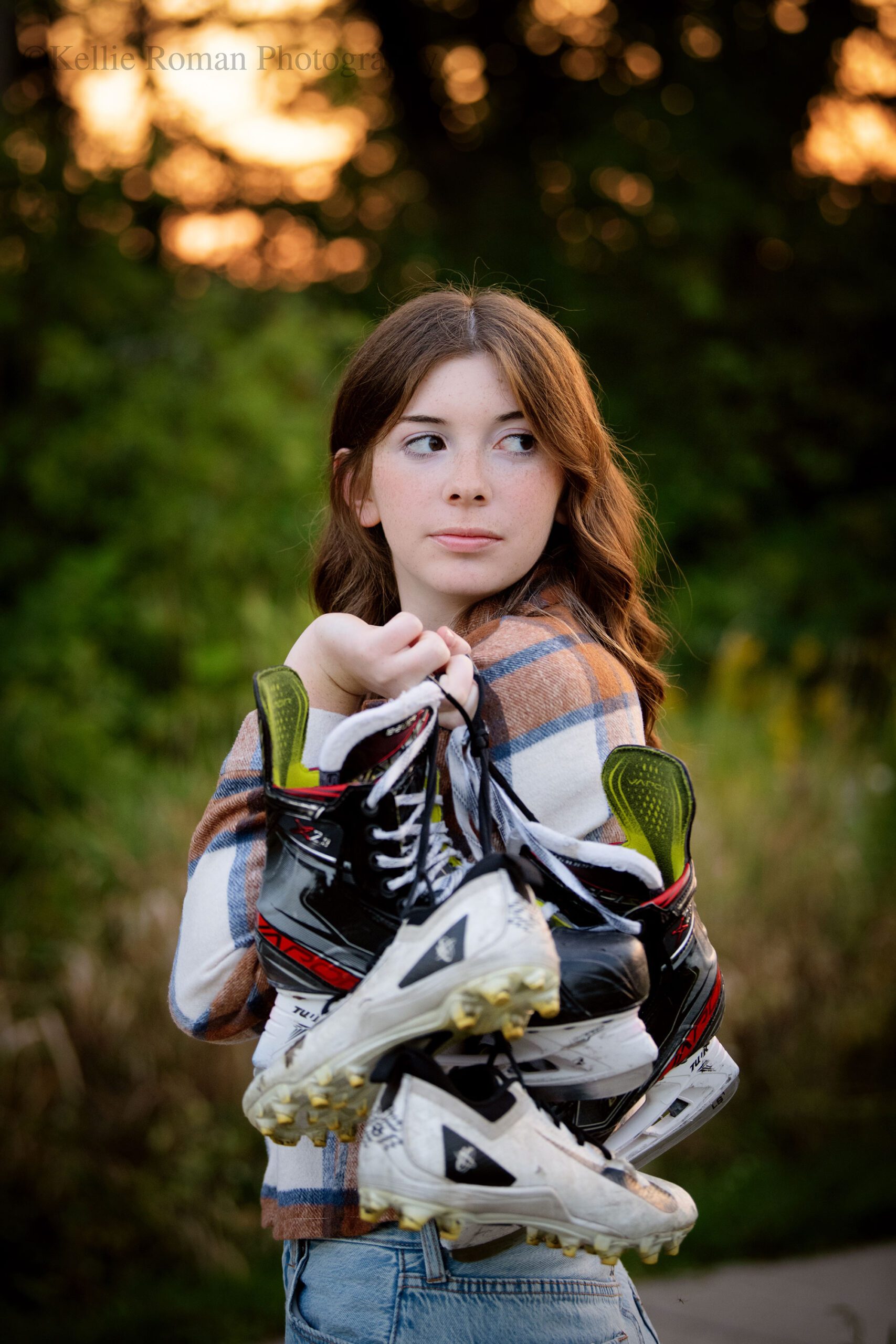 senior photography in milwaukee. high school senior girl wearing jeans and flannel shirt has hockey skates and lacrosse cleats over her shoulder while she is looking behind her.
